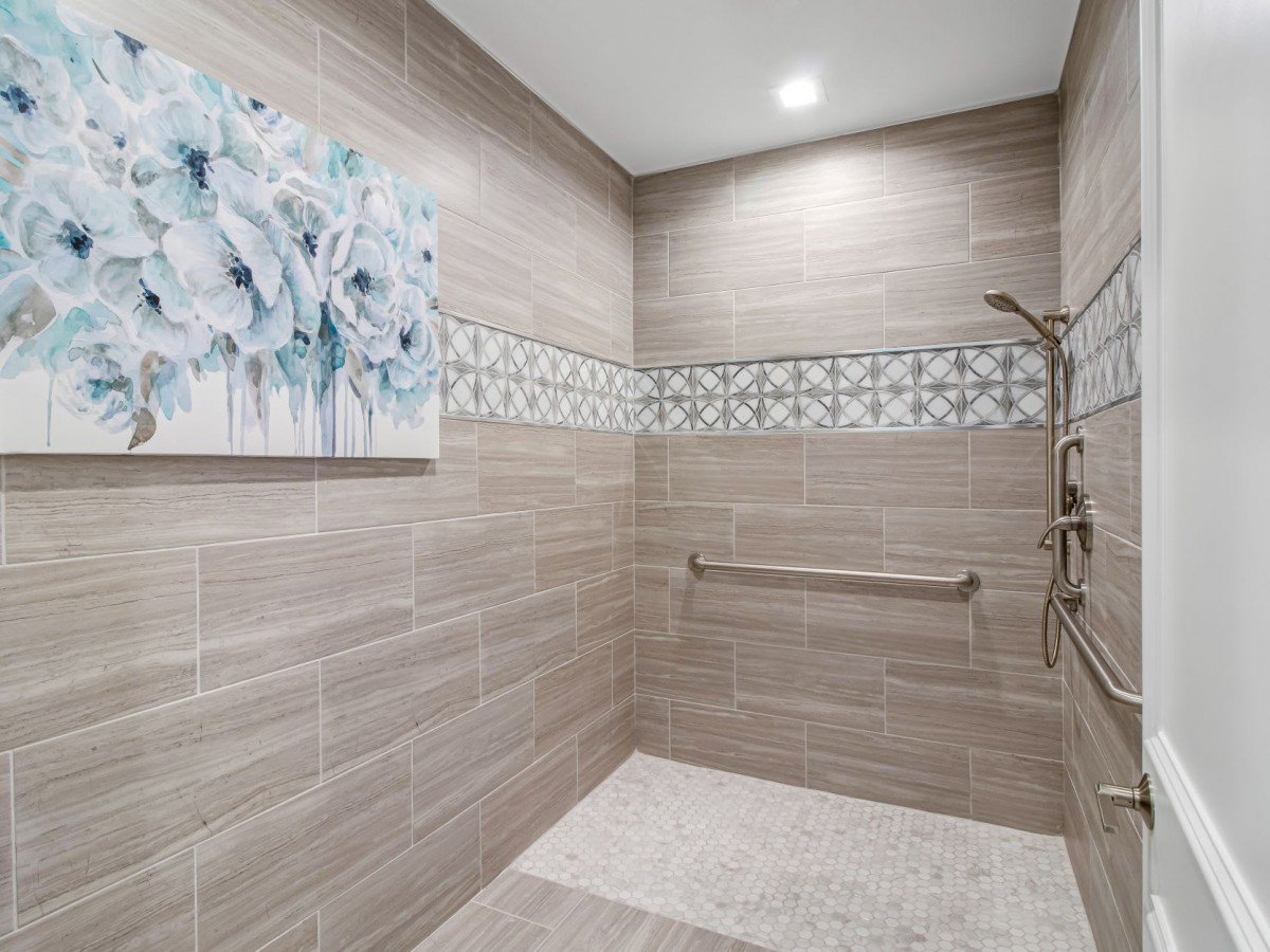 A modern shower with beige tiles and a decorative tile band, featuring a grab bar and wall artwork.