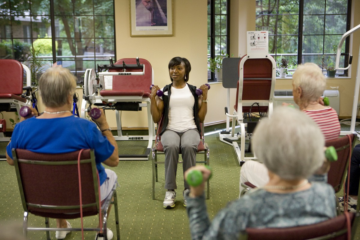 A fitness instructor leads a group of seniors in chair exercises using weights.