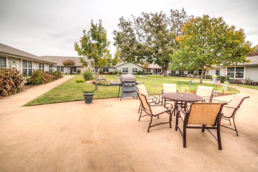 An inviting outdoor patio with a table, chairs, and grill set against a backdrop of a well-maintained lawn and trees.