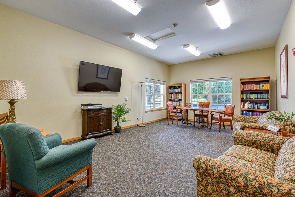 A cozy living room with a television, bookshelves, chairs, and large windows.