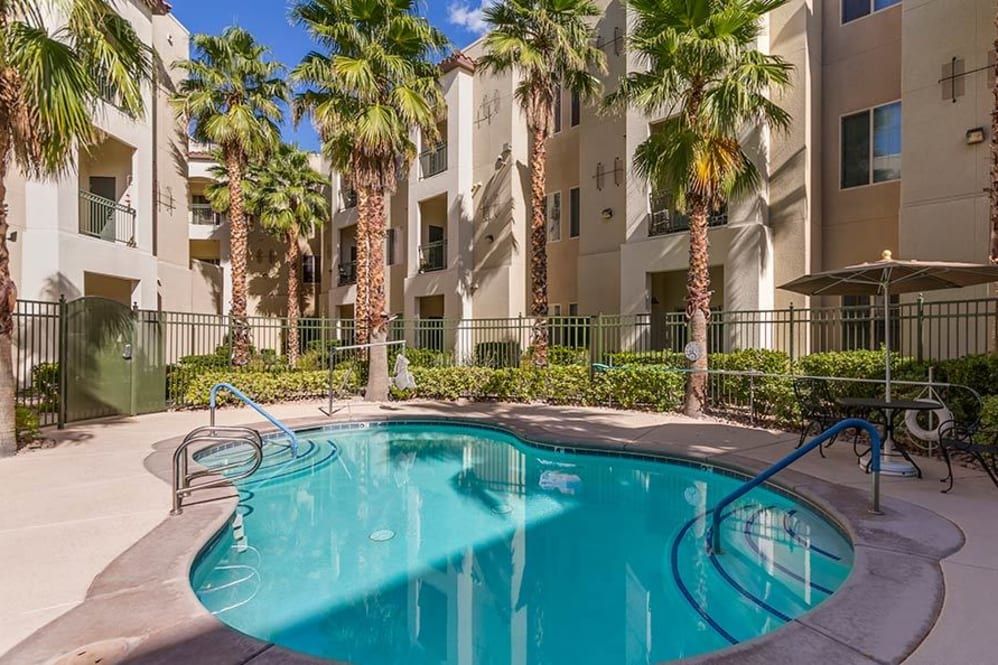 A serene outdoor swimming pool area surrounded by palm trees and a residential apartment complex on a sunny day.