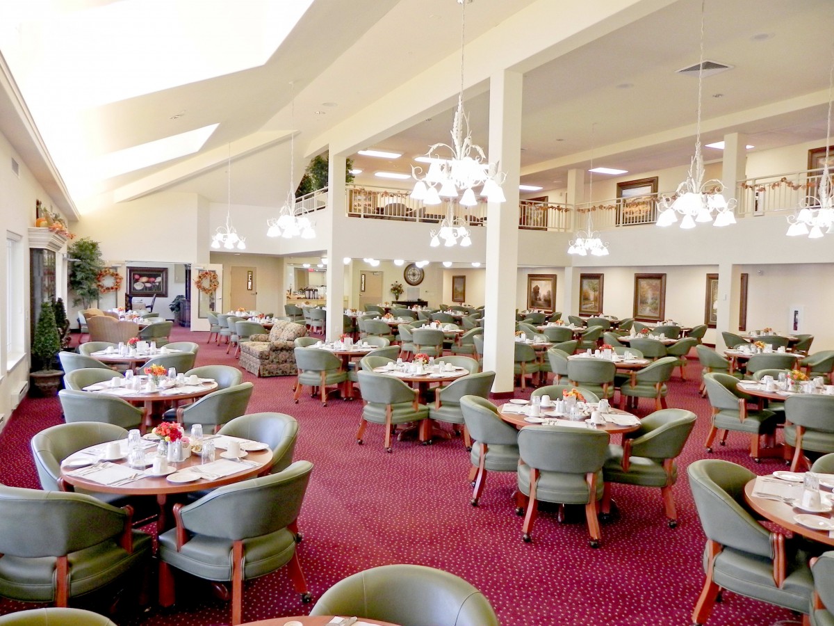 A spacious dining area with elegant chandeliers, neatly arranged tables and chairs, and a vibrant red carpet.