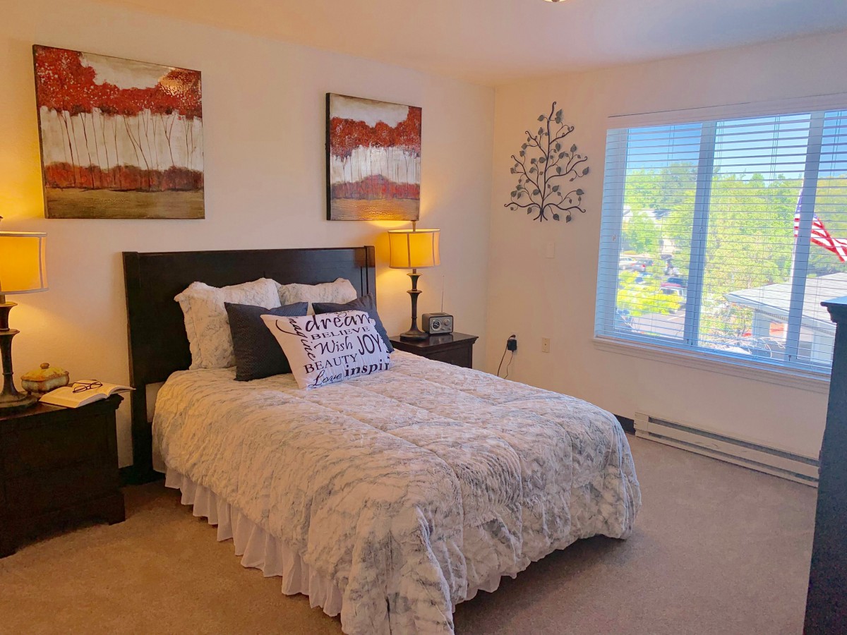 A cozy bedroom featuring a bed with decorative pillows, bedside tables with lamps, and wall art under natural light from a window.