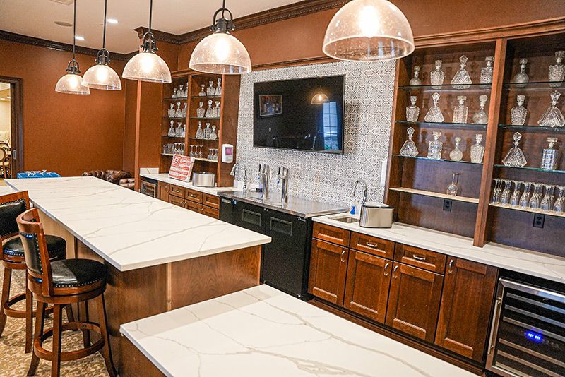 A modern kitchen interior featuring wood cabinets, elegant countertops, shelving units, pendant lights, and a large TV screen.