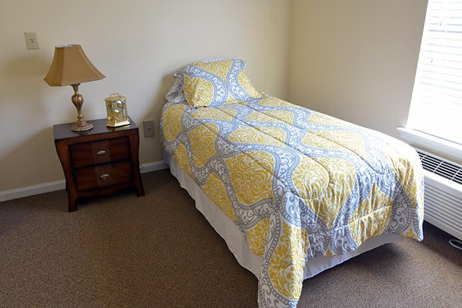 A cozy bedroom featuring a single bed with a yellow and blue quilt, next to a wooden nightstand with a lamp.