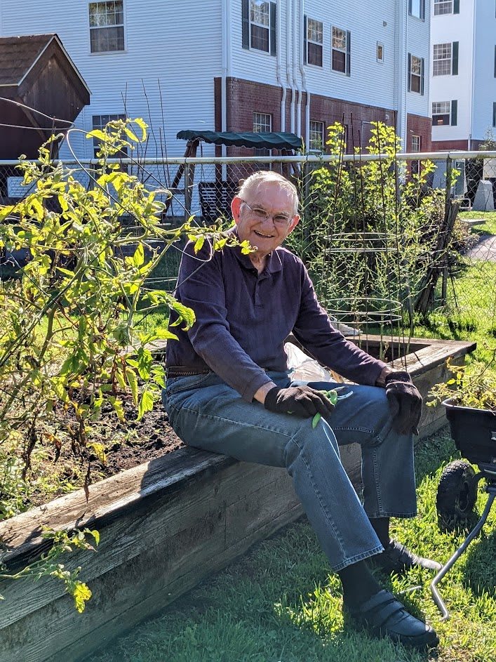 An elderly man enjoying a sunny day while gardening, seated near a wheelbarrow.