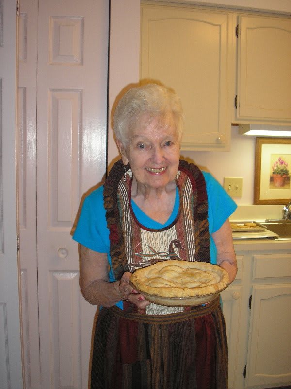 An elderly woman smiling and holding a homemade pie in her kitchen.