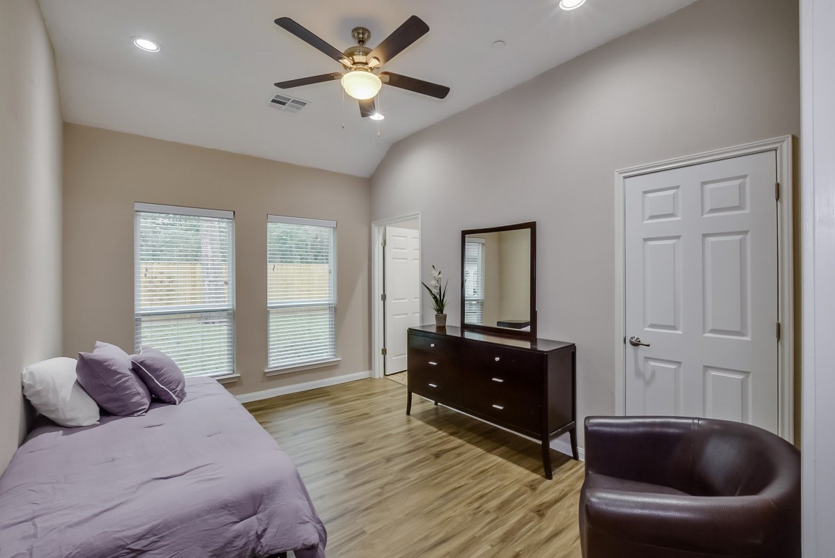 A modern bedroom with wooden flooring, featuring a bed, dresser with mirror, ceiling fan, and large windows.