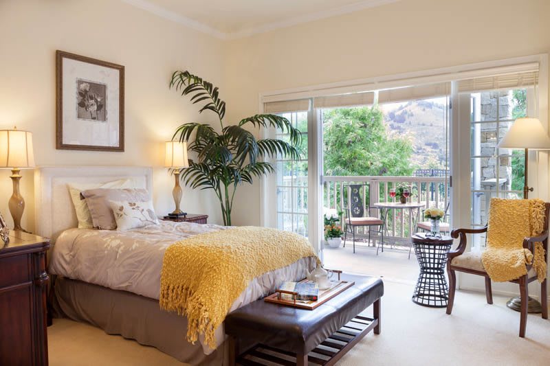 A cozy and elegantly decorated bedroom with a large window leading to a balcony, allowing natural light to fill the space.