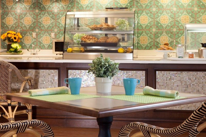 A cozy cafe interior with a wooden table set for two, featuring blue cups and a potted plant. The background showcases a display counter and elegant green patterned wallpaper.