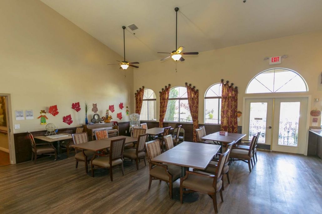 A spacious dining area with multiple tables and chairs, featuring large windows, ceiling fans, and decorative elements on the walls.