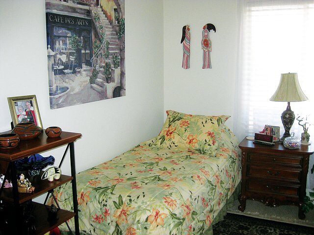 A small, cozy bedroom with a floral bedspread, wall art, and a nightstand.