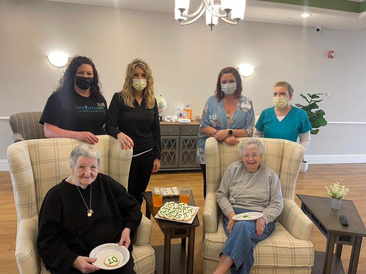 A group of elderly residents seated with caregivers standing behind them in a comfortable indoor setting.
