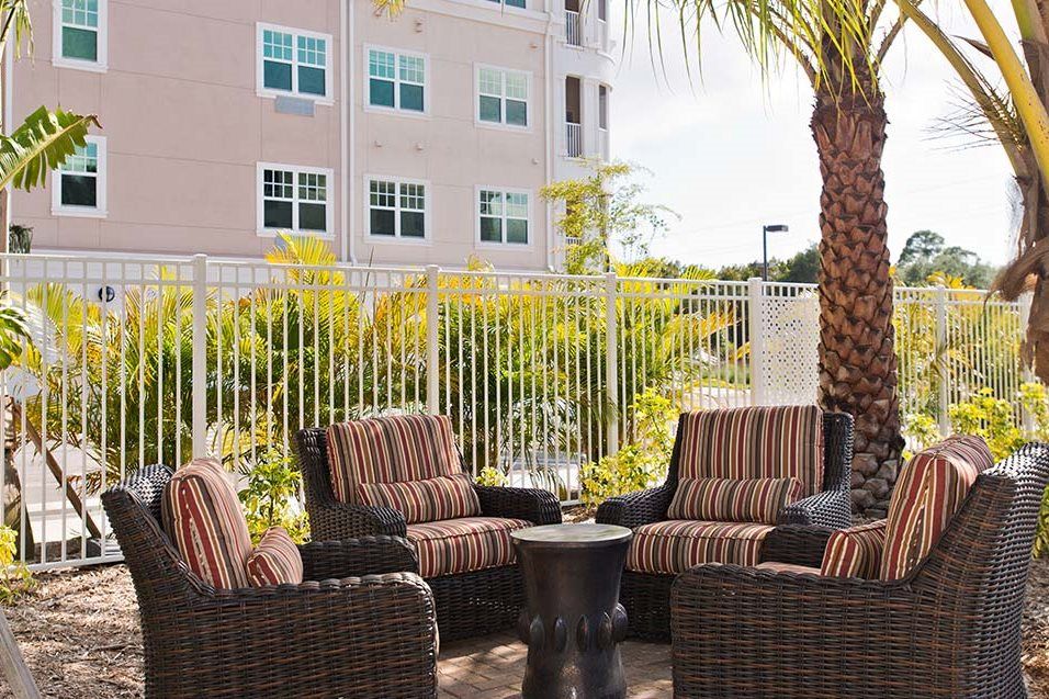 A cozy outdoor patio area with wicker chairs and a small table, surrounded by a palm tree and fence.
