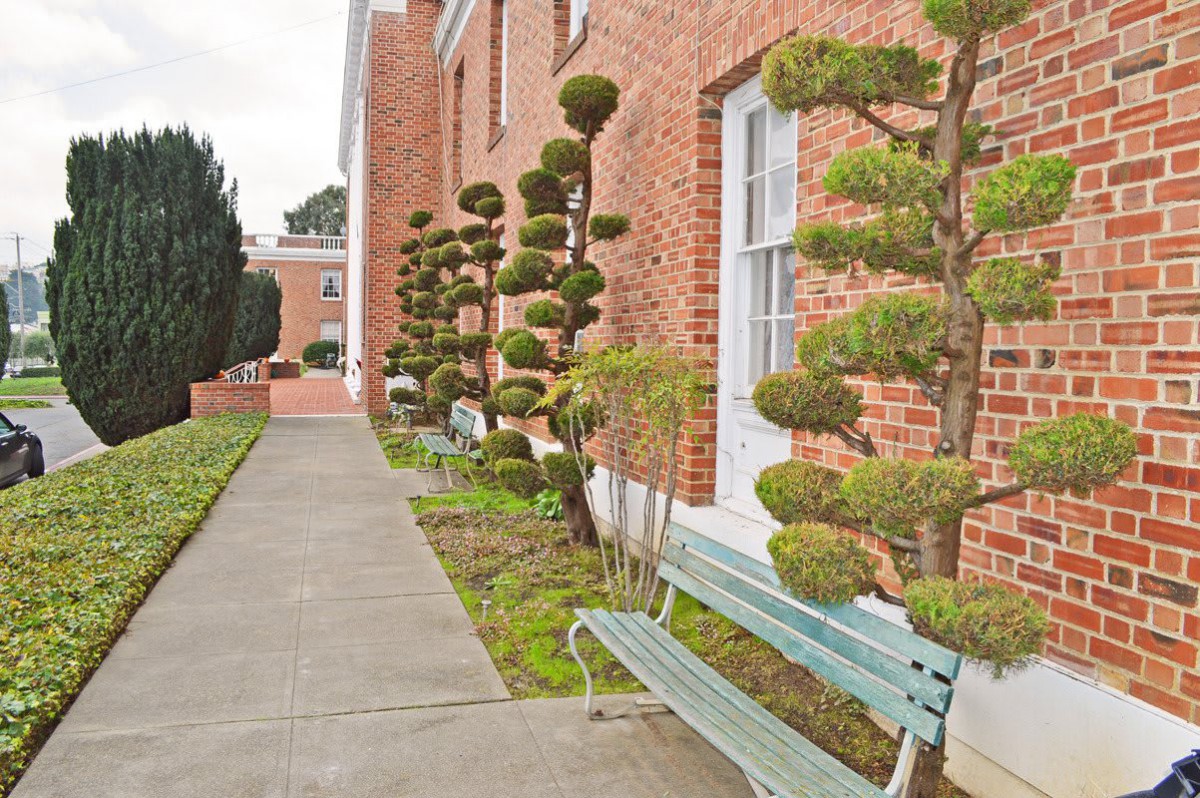 A walkway beside a brick building features uniquely shaped topiary plants and a green bench.