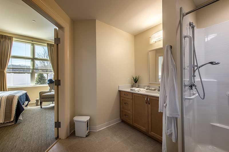 A well-lit bathroom with a wooden cabinet, sink, and mirror adjacent to a bright room.