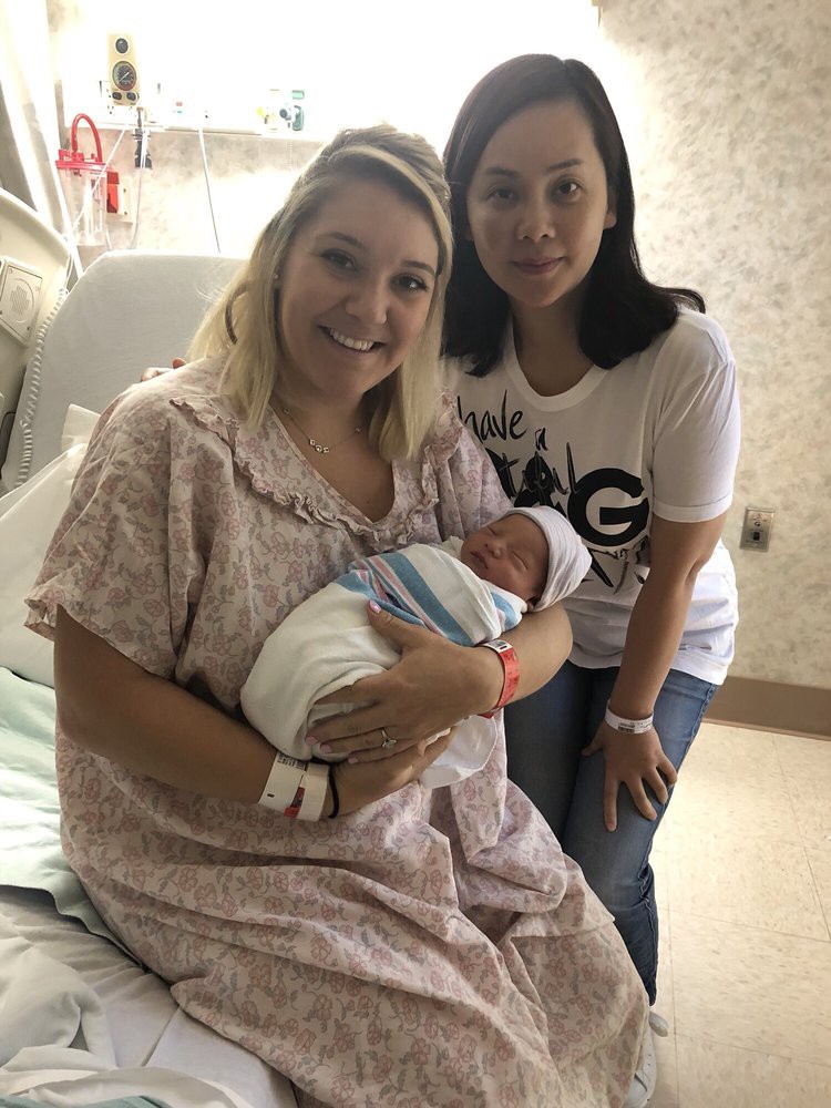 A mother holding her newborn in a hospital room with another person standing beside them.