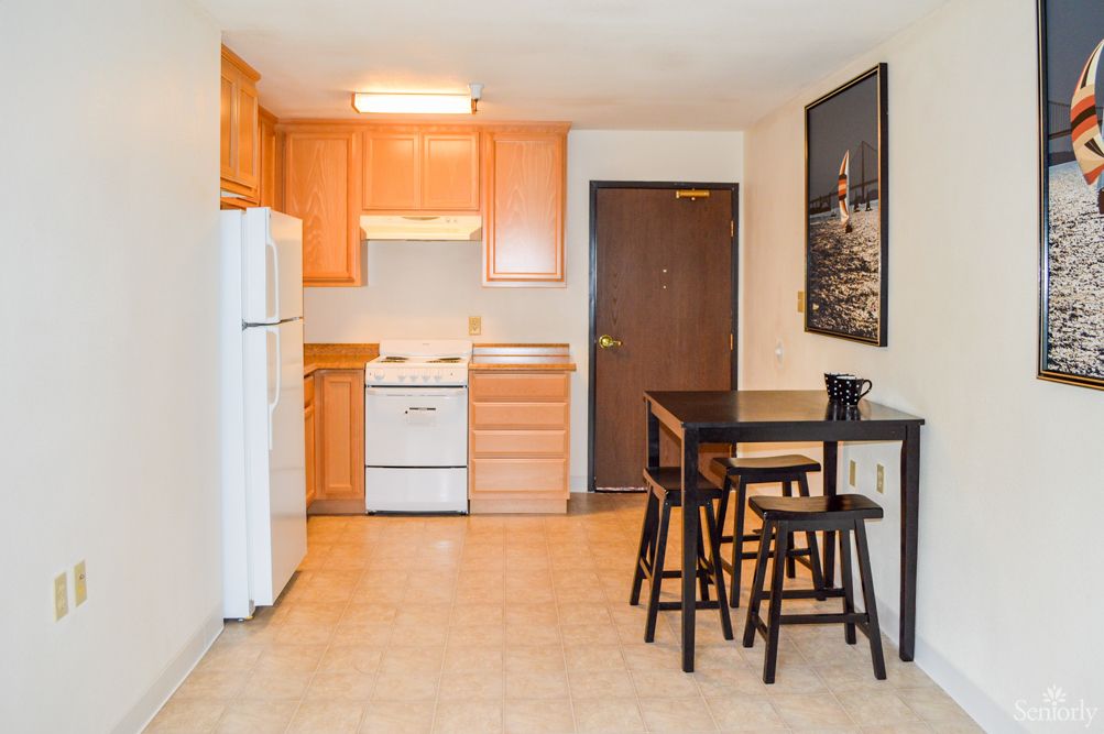 A compact apartment kitchen with wooden cabinets and a small dining area featuring a table and stools.