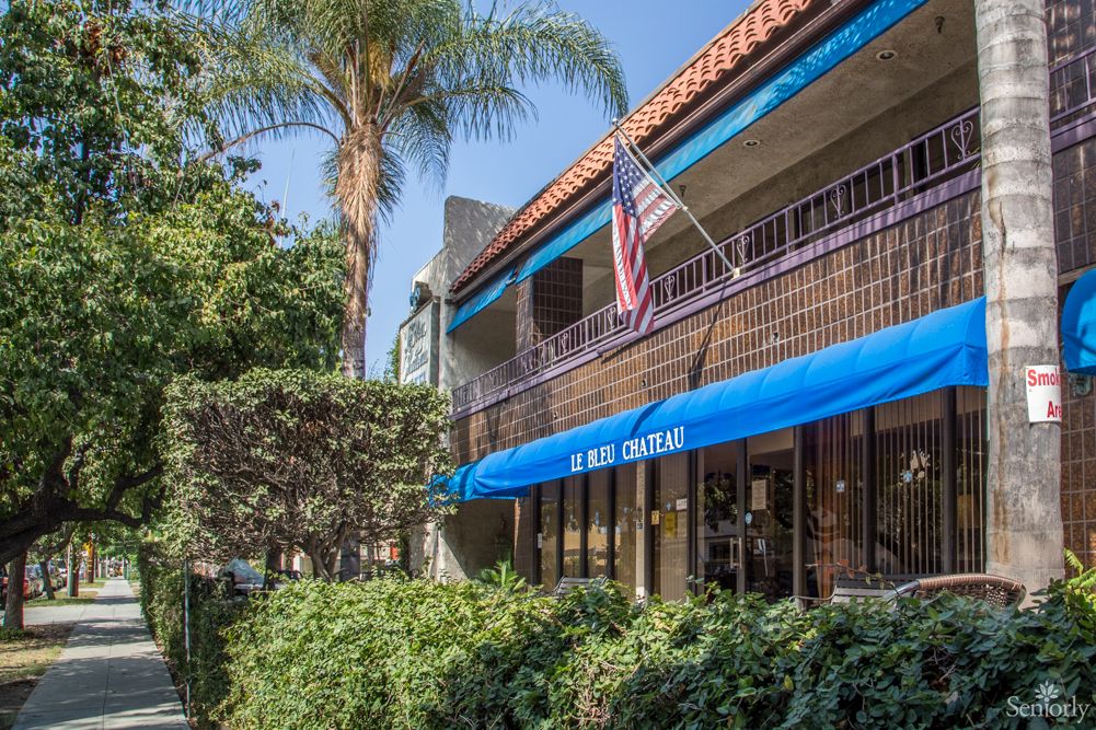 An outdoor view of a building featuring a blue awning and an American flag, surrounded by lush greenery and palm trees.