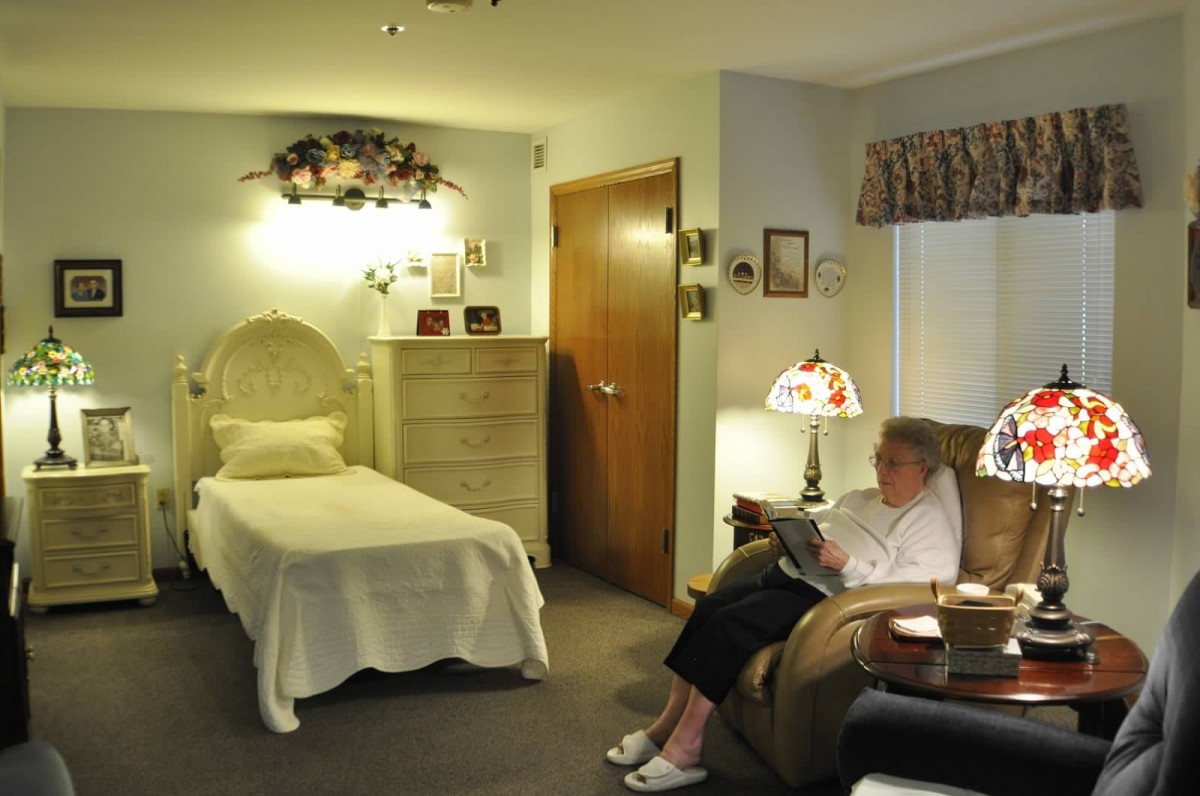 An elderly woman sitting and reading in a cozy bedroom with a lamp.