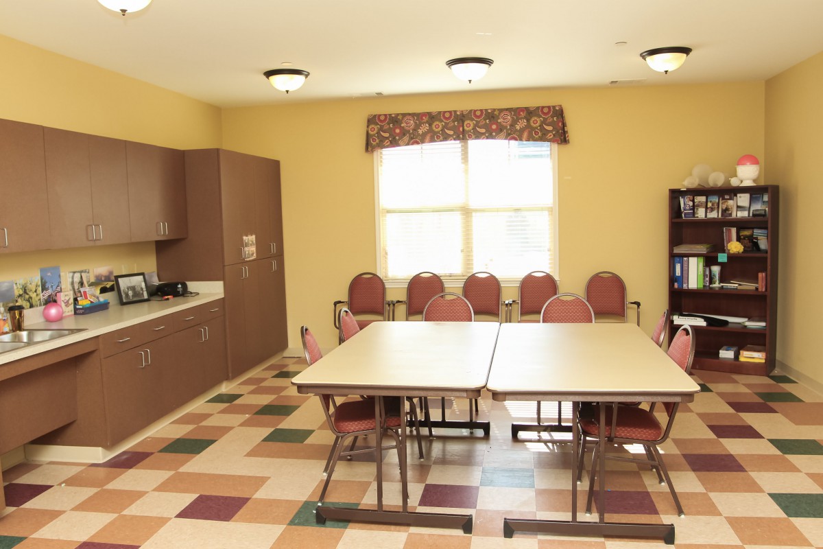 A small meeting room with tables and chairs, featuring a kitchenette and large window.