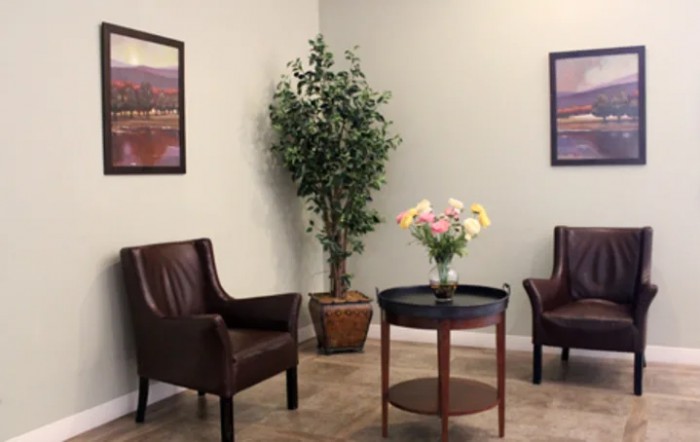 A cozy waiting room with leather chairs, a round table adorned with flowers, a potted plant in the corner, and framed paintings on the wall.