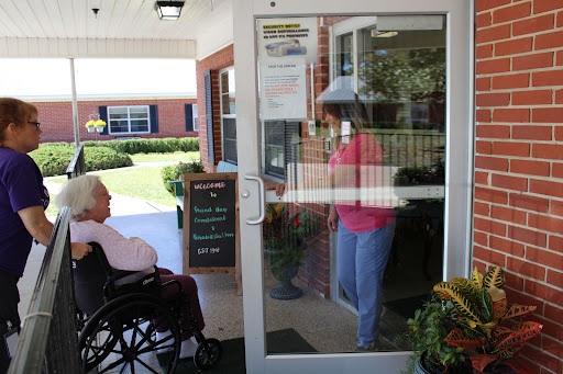 A senior person sitting in a wheelchair while another individual approaches the door of a care facility.