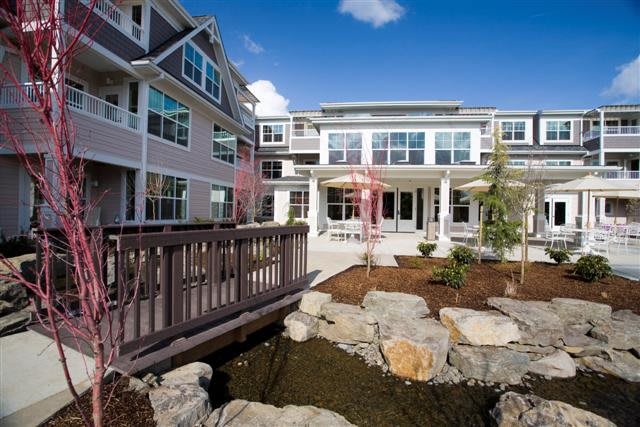 A modern residential building with landscaped surroundings, featuring a small bridge over a pond.