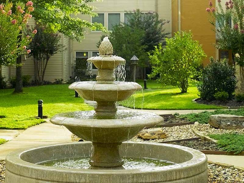 A multi-tiered fountain in a garden setting with lush greenery and a building in the background.