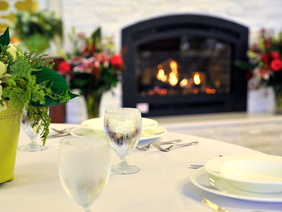 A cozy dining setup featuring a table elegantly set with glassware and surrounded by floral arrangements, with a warm fireplace glowing in the background.