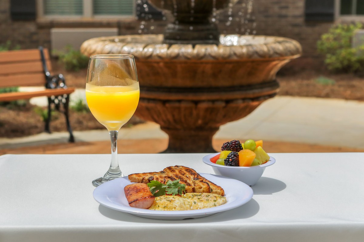 A delightful breakfast featuring grilled chicken with herbs, a fresh fruit bowl, and a glass of orange juice served at an outdoor table with a fountain backdrop.