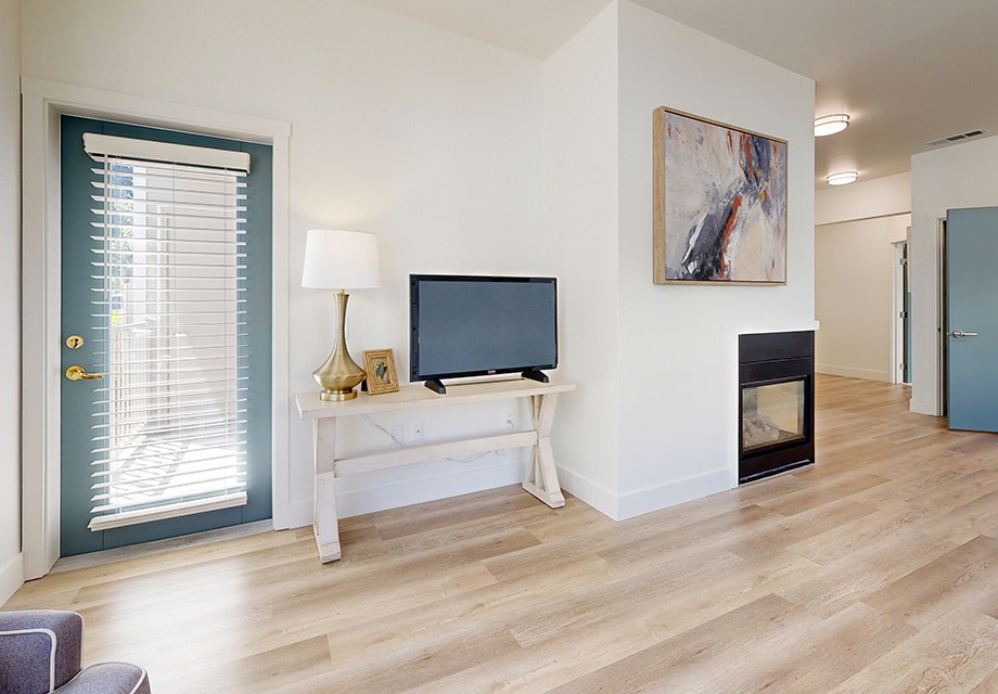 A minimalist living room featuring a TV on a white console table, an abstract artwork above a modern fireplace, and light wood flooring.