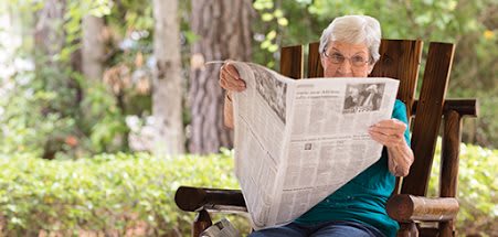 An elderly person sits on a bench outdoors, reading a newspaper.