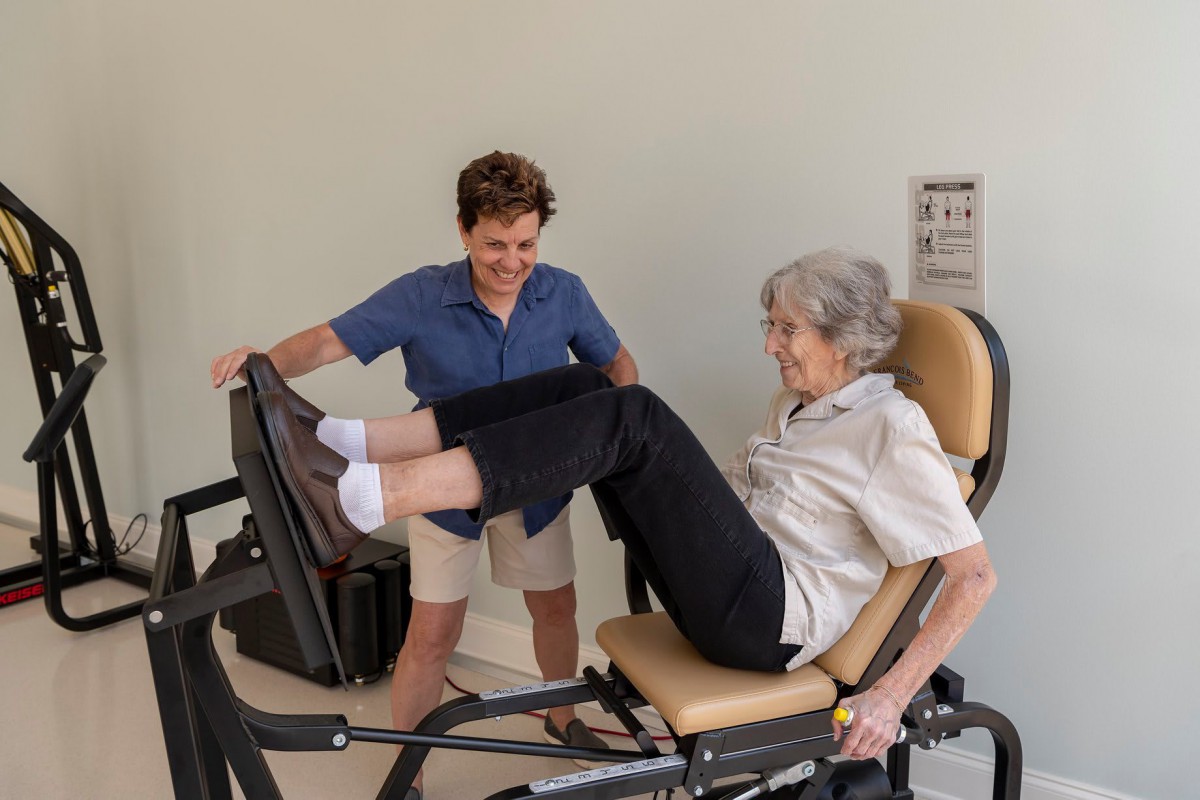 An elderly woman exercises on a leg press machine with the assistance of a trainer.