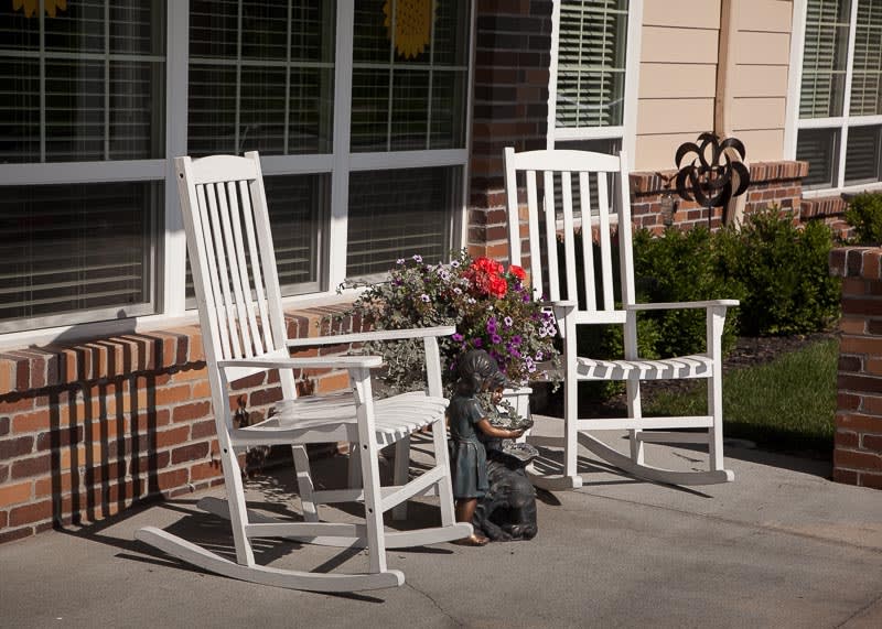 Two white rocking chairs on a sunny porch with flowers and plants.