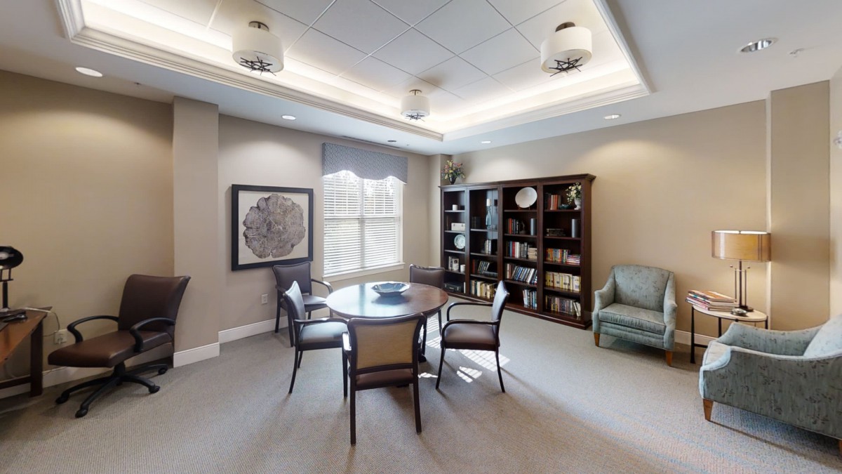A cozy office space featuring a round table surrounded by chairs, with bookshelves and large windows providing ample natural light.