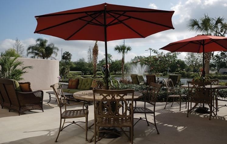 A tranquil patio setting with a table and chairs under a red umbrella, overlooking a garden and fountain.