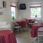 A small cafeteria with red tablecloths and foldable chairs, featuring a wall-mounted TV and windows.