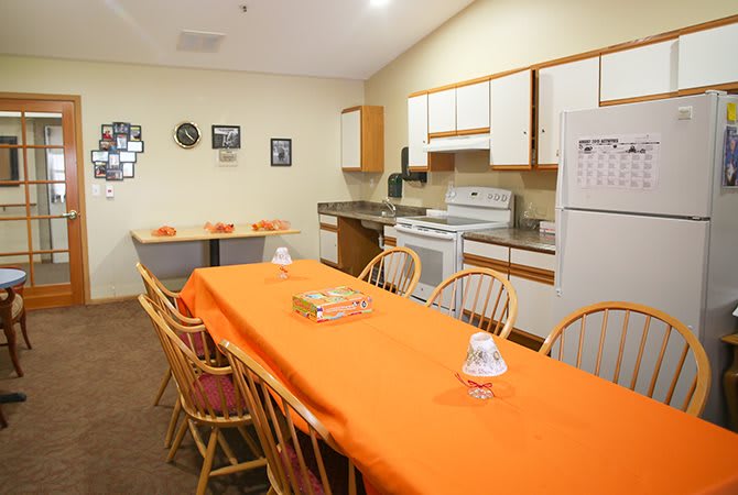 A kitchen and dining area with orange table settings, featuring a stove, refrigerator, and wooden chairs.