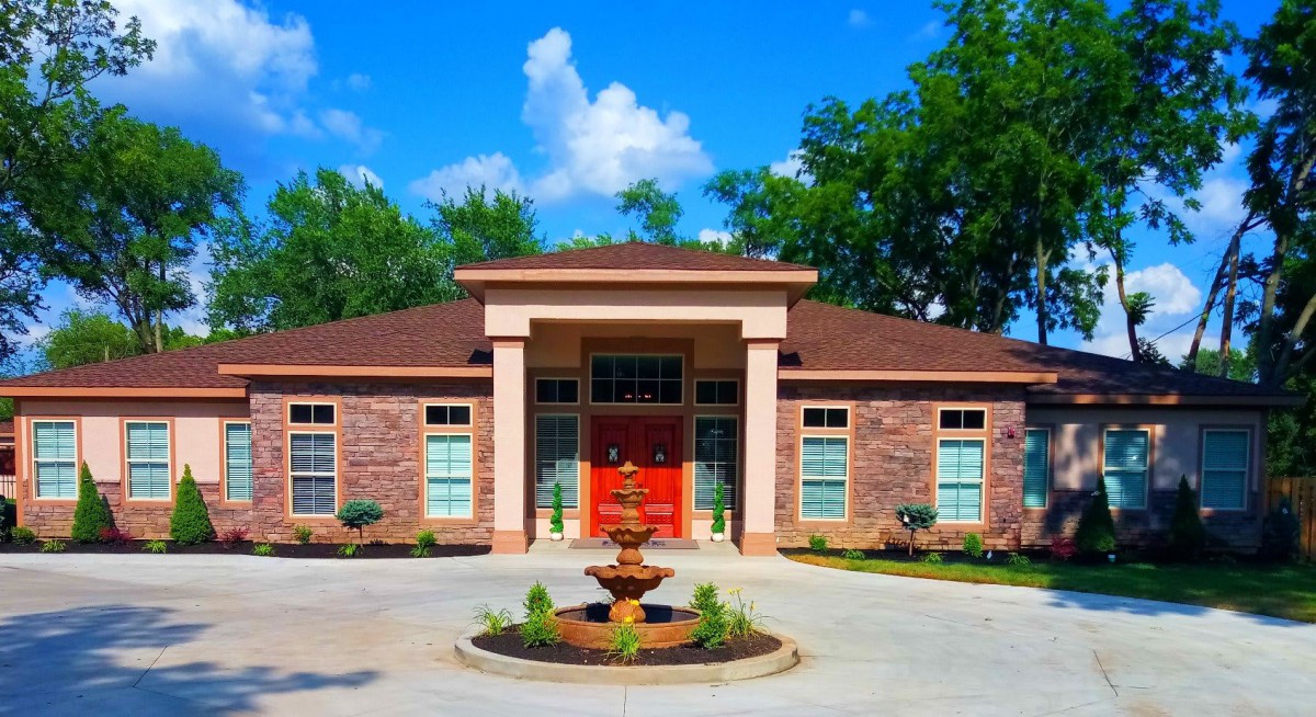 A stylish brick house with a red door and a central fountain, surrounded by greenery under a clear blue sky.