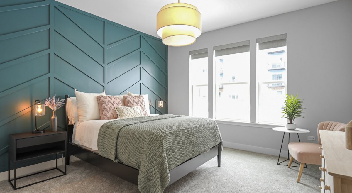 A modern bedroom with a blue accent wall, featuring a neatly made bed with decorative pillows and a green blanket. Ample natural light streams through large windows.