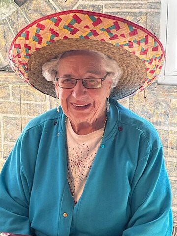 An elderly woman smiling warmly while wearing a sombrero indoors.