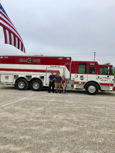 A group posing in front of a fire truck, with an American flag waving in the background.