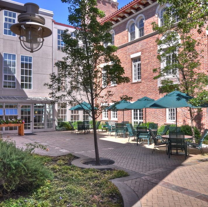 A charming urban courtyard with outdoor seating, tables, and umbrellas surrounded by a brick building and lush greenery.