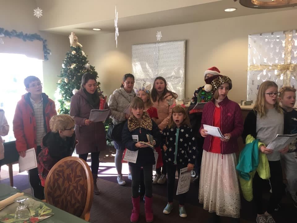 A group of people, including children, are gathered indoors singing Christmas carols in a festive setting.