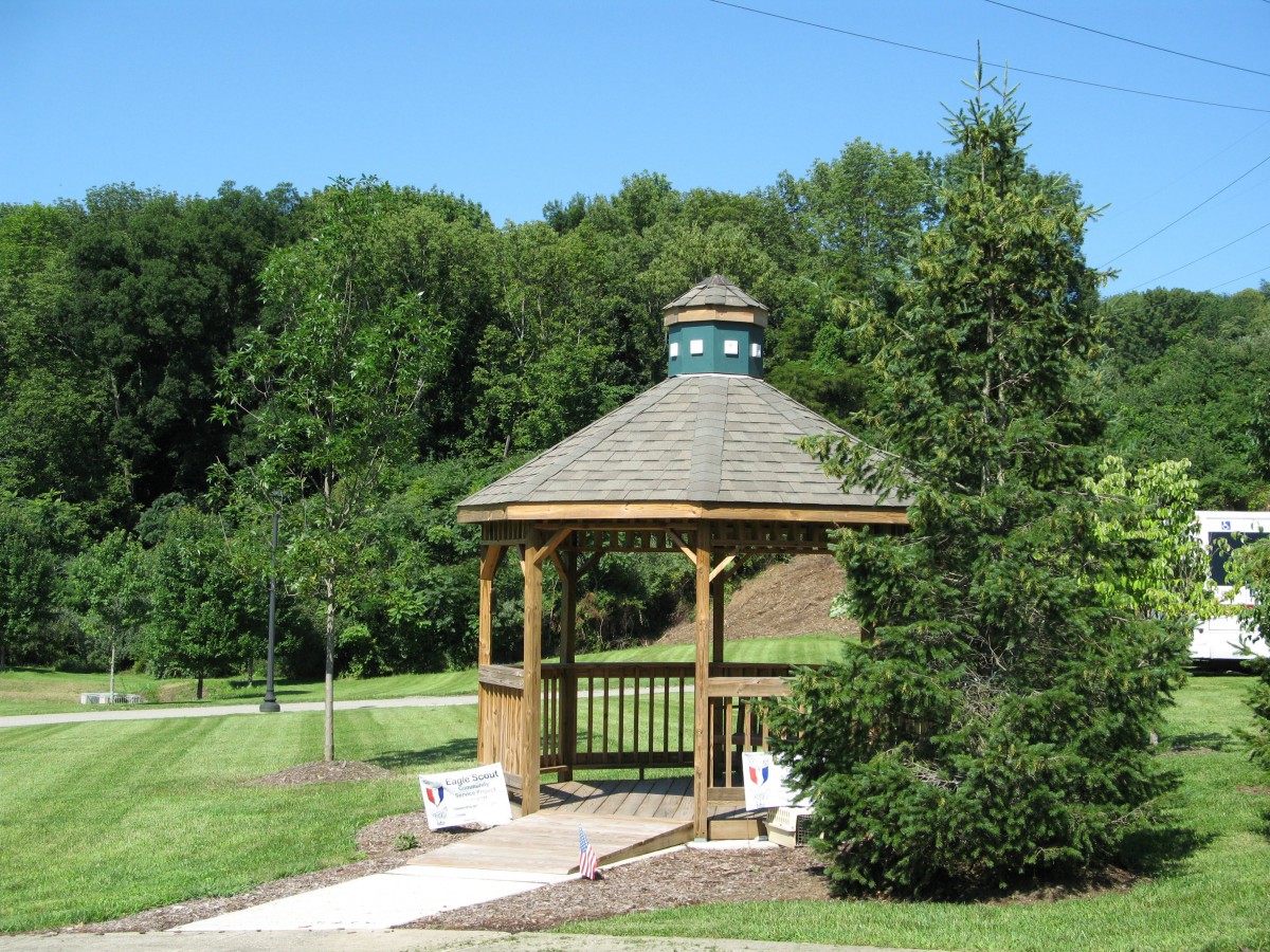 A wooden gazebo surrounded by greenery in a park setting with benches and a pathway.