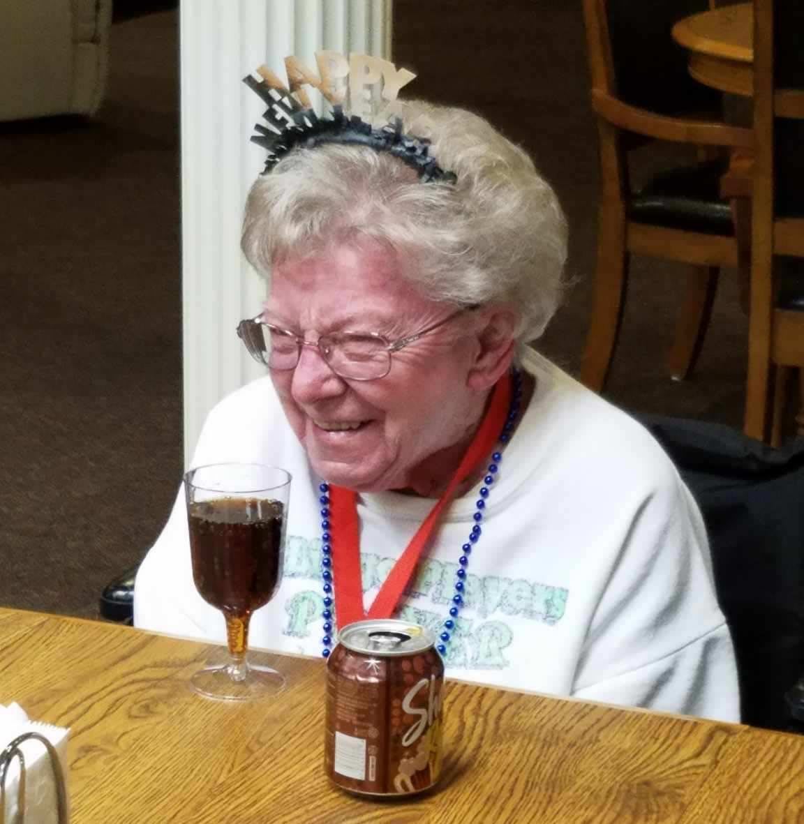 An elderly woman is smiling and wearing a crown at a table with a soda can and glass.
