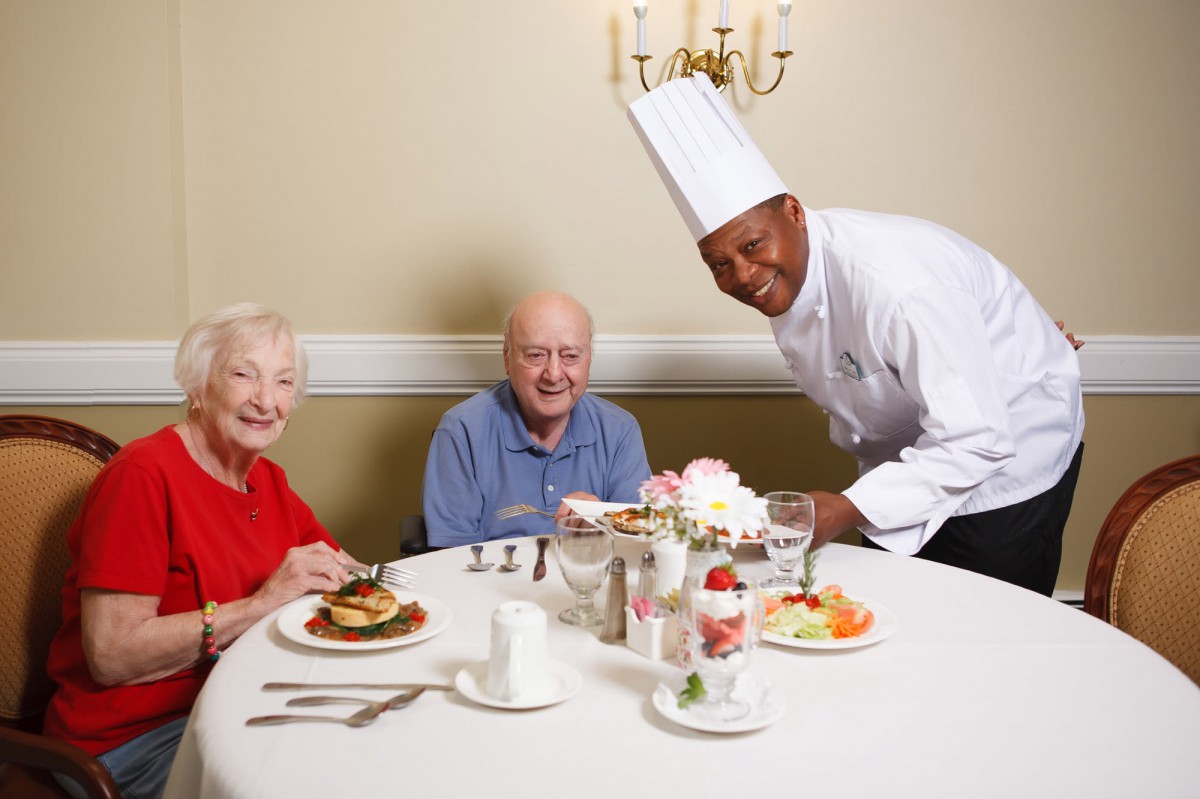 An elderly couple enjoying a meal served by a chef in a restaurant setting.