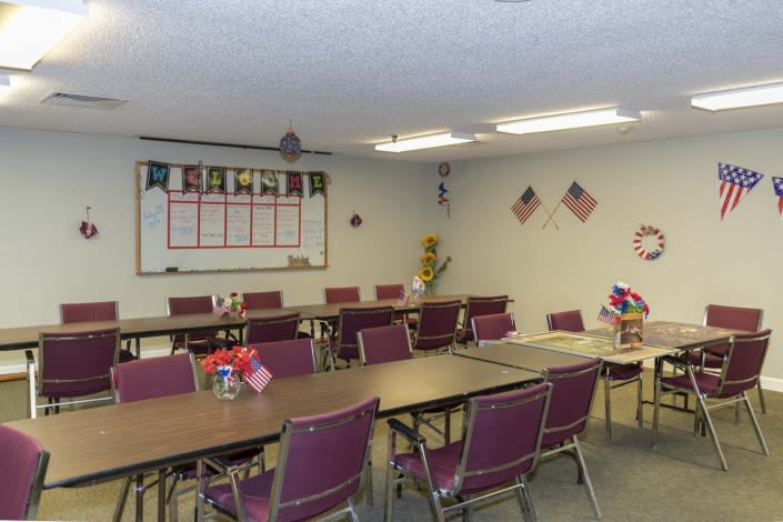 An empty classroom with tables and chairs arranged in rows, a whiteboard on the wall, and various decorations.