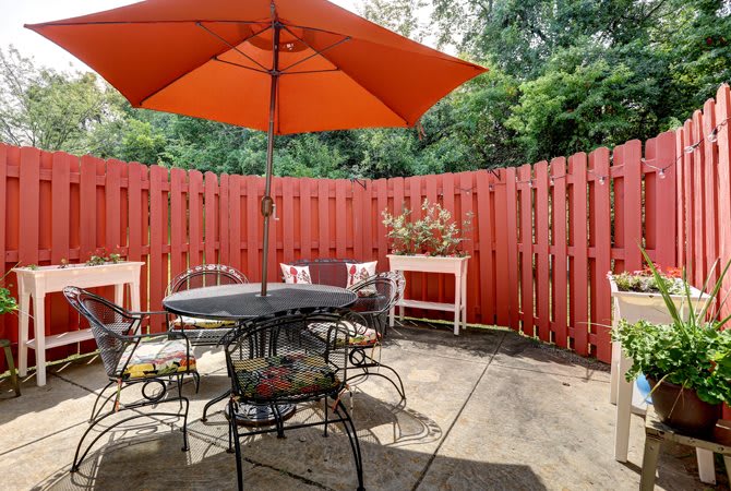A cozy outdoor patio space with a red fence, featuring a table and chairs under an orange umbrella.